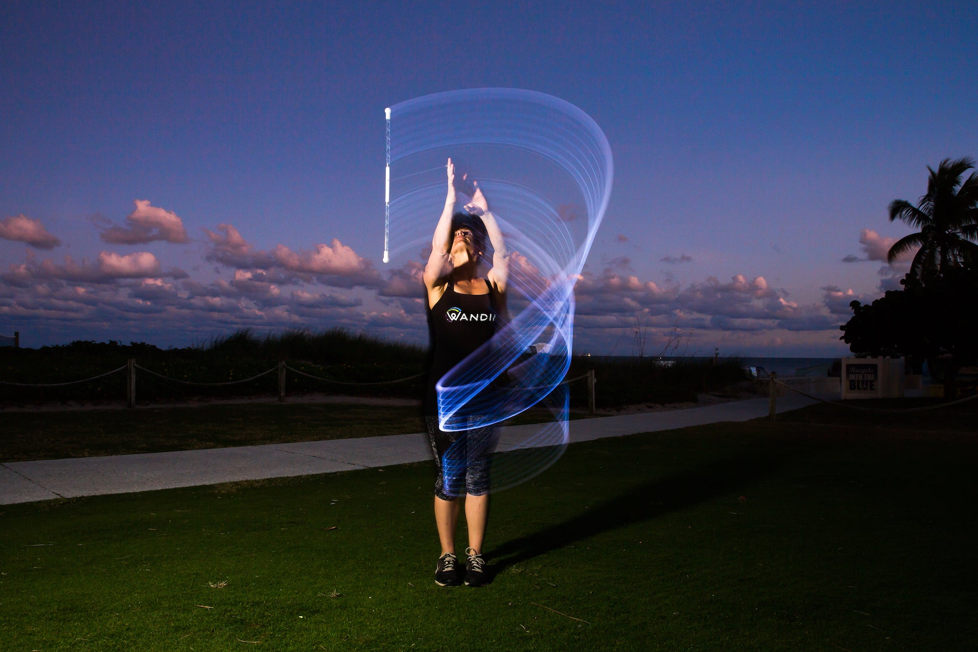 A performer using the Wandini Glow.0 Magic LED Levitation Wand outdoors at dusk, creating a glowing light trail in the air with the wand.